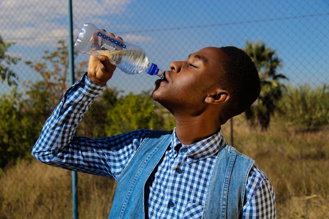 Side profile of a young man enjoying water from a bottle in a sunny outdoor setting.